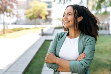 Portrait of pleased beautiful hispanic or brazilian curly haired young woman, in stylish clothes, successful mixed race businesswoman, standing outdoors with arms crossed, looking away, smiling