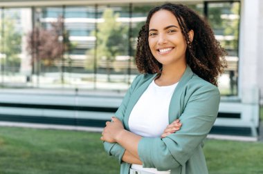 Portrait of successful pretty hispanic or brazilian curly haired young stylish woman, mixed race confident business woman, standing outdoors with arms crossed, looking at camera, smiling friendly