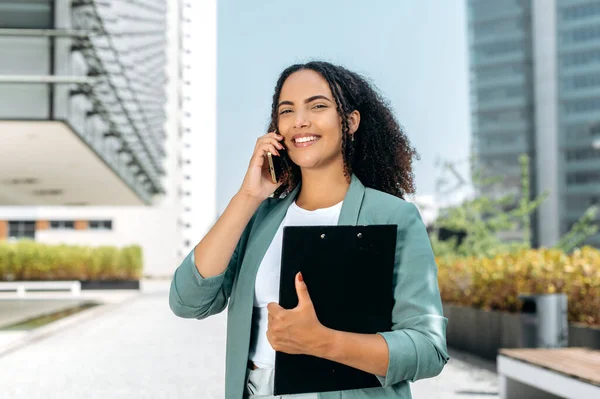 Cell Phone conversation. Confident positive young mixed race curly successful business woman, real estate agent, in elegant clothes, has conversation on a smartphone while standing outdoors, smiling