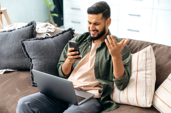 Positive mixed race young man in casual clothes, spending time at home, sits on the sofa in the living room, using a smartphone, read unexpected news, browsing the internet, social networks, smile