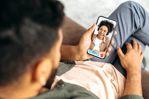 Online video consultation. On a screen of the phone, an African American lovely positive girl holds consultation. Husband and wife or friends talk on online video conference using mobile phone, smile