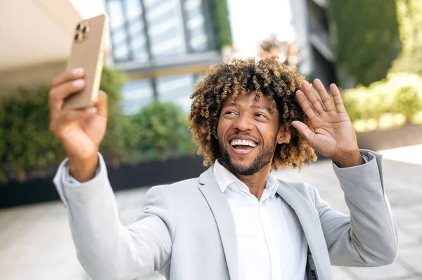 Positive handsome curly haired hispanic or brazilian man standing outdoors, taking a selfie on her smartphone, mixed race male blogger having live streaming on social network, smiling. Video call