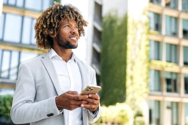 Online messaging. Positive handsome confident curly haired brazilian or hispanic man, successful entrepreneur, standing outdoors, using smartphone, messaging in social media, looking away, smile