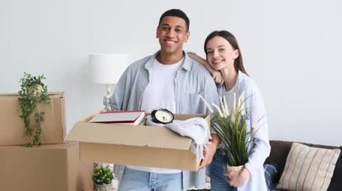 Positive happy couple in love, husband and wife of different nationalities, moved to their new home, the guy holds a box with things, the girl keys and a flowerpot, look at the camera, smile