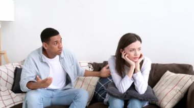 Misunderstanding in relationships, jealousy, quarrel. Mixed race couple sitting on a sofa in the living room, an angry man yells at a woman, feels angry, frustrated sad girl turned away from him