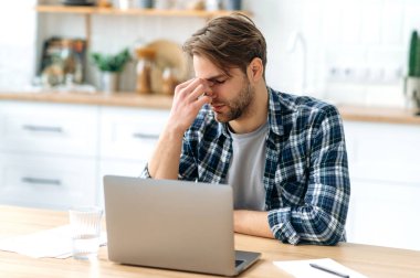Frustrated sad caucasian man, closed eyes, massages his bridge nose, sitting at his workplace at home office in the kitchen, take break. Feeling exhausted and tired during long work in the laptop