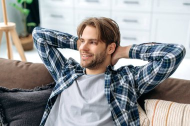 Close-up of calm relaxed satisfied caucasian guy in casual shirt, resting on a comfortable sofa in cozy living room, throwing his hands behind his head, dreaming of vacation, resting from work, smiles