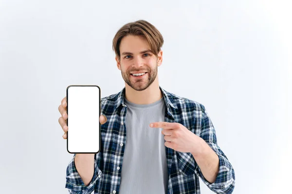 Attractive positive caucasian young man, looks at camera, hold in hand smartphone with white empty mock-up screen for presentation and points finger at it, stand on isolated white background, smiles