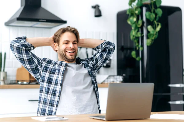 Calm satisfied caucasian guy, freelancer, working from home, sits in the kitchen at workplace, takes a break from work, puts his hands behind his head, looks to the screen, glad with a project, smiles