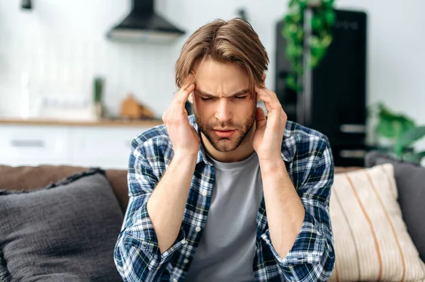 Close-up of upset exhausted caucasian man, sits on a sofa in the living room, massages his temples, closed his eyes, feels unwell, suffering migraine, fatigue, needs treatment, rest. Headache, spasm