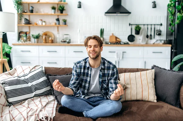 Calm happy peaceful relaxed caucasian young man in casual clothes, sitting alone at home in living room on the couch and meditating in the lotus position, smiling. Relaxation and meditation concept