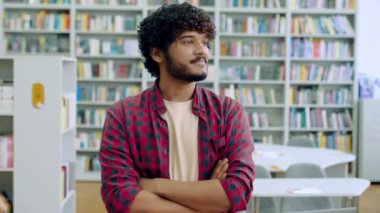 Yakışıklı, kendine güvenen kıvırcık ırklı adam, Hintli ya da Arap erkek, günlük giyim, öğrenci, serbest çalışan ya da öğretmen, kütüphanede arka planda duruyor, kameraya bakıyor, gülümsüyor.