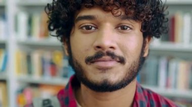 Close-up video of a handsome positive curly haired indian or arabian guy, in casual shirt, manager, designer, student or freelancer, stands indoors, looks at camera, smiling friendly