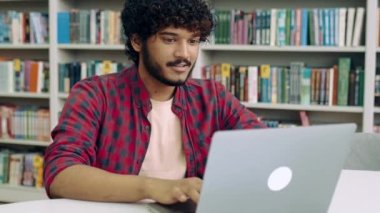 Relaxed calm satisfied mixed race student guy, freelancer, sits in the library at a desk with laptop, takes a break from study, puts his hands behind his head, looks away, glad with a project, smiles