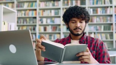 Positive smart arabian or indian college male student, sits at table with laptop in university library, preparing for exam, studying information, rereading his abstract, gain knowledge, smiles
