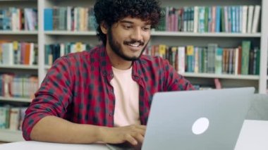 Satisfied clever arabian or indian student guy, freelancer, sits in the library at a desk with laptop, takes a break from study, puts his hands behind his head, looks away, glad with a project, smiles