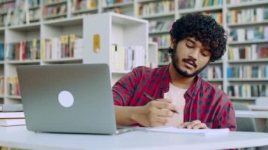 Focused clever arabian or indian college student, sits at table with laptop in university library, preparing for exam, studying information, listening lecture, taking notes in notebook, gain knowledge
