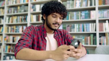 Happy indian or arabian millennial guy, freelancer or student, using his smartphone, chatting with friends on social networks, texting sms, browses internet,smile friendly. Wireless technology concept