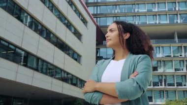 Successful elegant confident young mixed race woman with curly hair, formally dressed, stands outdoors against the background of the business center, with arms crossed, looks at camera, smile friendly