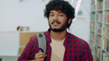 Positive clever curly haired mixed race male student of university, in stylish clothes, with backpack, walks across the library near the background of bookshelves, looks at camera, smiles friendly