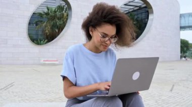 Beautiful positive confident African American curly haired young woman, with glasses, manager, company ceo, freelancer, sits with laptop outdoors near business center, work online, smiles friendly