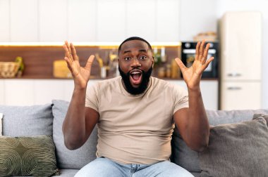 Cheerful happy young african american man, sitting on the sofa in living room, excited for success with arms raised, celebrating victory, goal, smiling happily, screaming, looks at camera in amazement