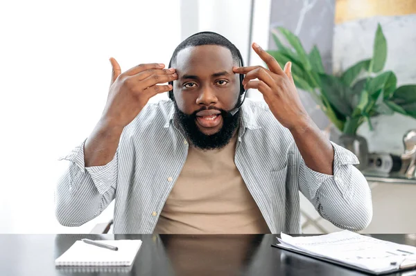 Stunned puzzled african american man with headset, business leader or coach, talking with colleagues by video call, discuss ideas and promotion, looks at camera in confusion, gesturing hands