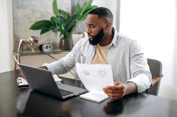 Focused busy successful african american young man, freelancer or company ceo, executive, works with documents, sit at desk in modern office, works on a project uses laptop, studying financial graphs