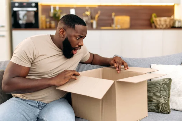 Shocked confused african american guy opening big carton box, unpacking internet store order at home, feels frustrated with long delivery service and the goods which received, experiencing puzzling