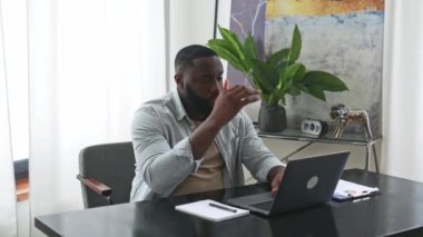 Sad exhausted african american young man, company employee, sitting at a desk, looks tired and stressed, experience headache, migraine, needs rest, closed his eyes. Overworked from work, deadline