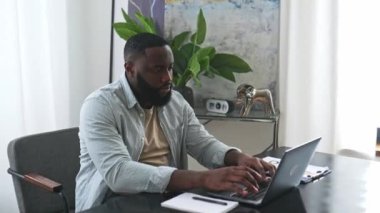 Attractive positive african american stylish man, entrepreneur or freelancer, IT specialist, working in a laptop while sit in modern office, develop creative successful project, looks at camera, smile