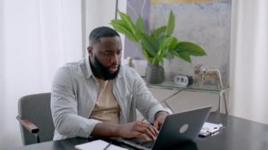 Frustrated sad stressed african american man, sit at desk, uses laptop for online work, annoyed with bad result of project, failed the deal, experiences negative emotions, stress,holding hands on head