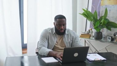Amazed happy emotional african american stylish man, trader, student, freelancer, sitting at table with laptop in the office, enjoying big profit, victory, good news, gesturing with fists, smiling