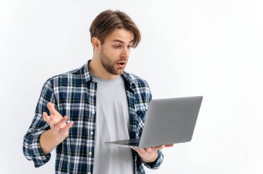 Shocked confused puzzled caucasian stylish young man, standing on a white isolated background, holds an open laptop, surprised looks at the screen, see unexpected news, message, gesturing with hand