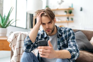 Sad confused upset caucasian young stylish man, sitting on a floor near a sofa in living room, frustrated looking at smartphone screen, failed project, got bad news or message, despair emotions