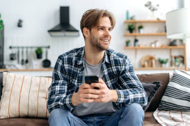 Positive handsome stylish caucasian young man, sitting with smartphone in hands on couch at home in living room, messaging online with friends, browsing websites, looks away, dreaming, smiling