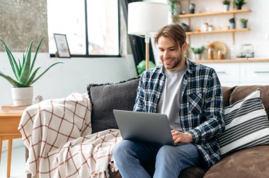 Joyful handsome stylish caucasian successful freelancer or designer, sitting with laptop on a sofa in modern living room while working or studying online, browsing information in internet, smiling