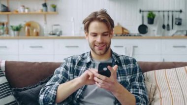 The concept of using gadgets. Attractive positive caucasian guy, sits in a modern living room on comfortable sofa, uses his smartphone, texting message, browsing social media, surfing web, smiles