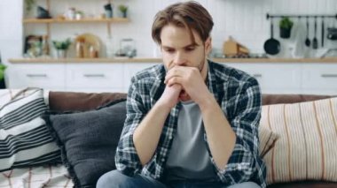 Sad depressed stressed caucasian man, sitting in the living room on the couch, looking ahead of himself, experiencing anxiety due to personal problems, unfulfilled expectations, something hurts