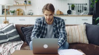Overworked stressed caucasian man in stylish clothes, freelancer, web designer, sitting in living room on the sofa, working on a laptop on a project, analyze documents, feels tired, suffering headache