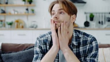 Close-up of upset exhausted caucasian man, sits on a sofa in the living room, feels unwell, suffering migraine, fatigue, experiences of personal troubles, feeling of anxiety