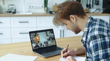 Online education. Side view of a smart caucasian male student and group of multiracial people on a laptop screen. Clever guy listening to an online lecture, takes notes, gain knowledge distantly