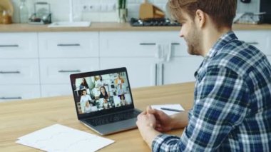 Side view of caucasian guy sitting in kitchen at home talking by video conference with group of multiracial successful colleagues discuss company promotion, advertising, financial strategy,new project