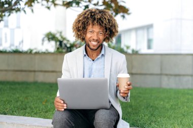 Positive attractive successful mixed race male freelancer, company worker, sitting outdoors, working online in laptop, taking break, holding glass of takeaway coffee, looking at camera, smiling