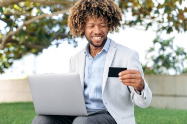 Positive handsome latino or brazilian man, sit outdoors, using a bank card and a laptop, making online purchases, making payments, ordering food delivery, enters data, smiles. Online shopping, payment