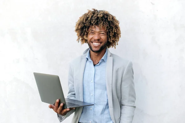 Portrait of handsome positive successful curly haired mixed race business man, elegantly dressed, holding open laptop in hand, stands on isolated background, looks at camera, smiling friendly