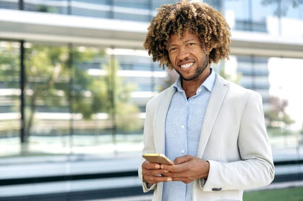 Online messaging. Successful confident elegant latino or brazilian curly haired man, wearing stylish formal clothes, stands outdoors near business center, uses his smartphone, looks at camera, smiles