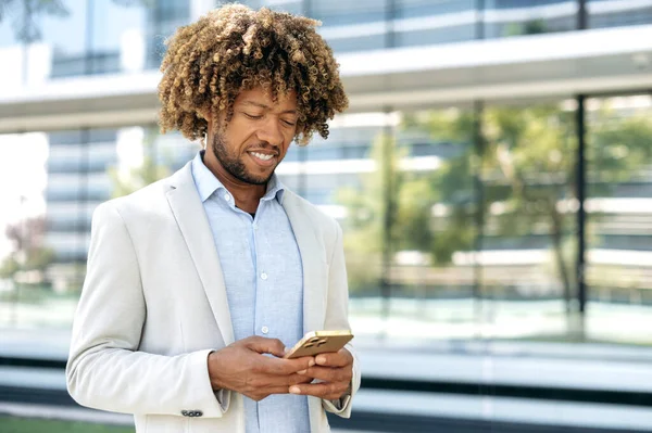 Online messaging, browsing internet. Positive mixed race curly haired man, wearing stylish formal clothes, stands outdoors near business center, uses his cellphone, looks at device screen, smiles