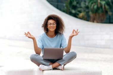 Peaceful calm relaxed african american young woman with glasses, office worker, sits in a lotus position outdoors near the business center, meditates, relaxes, closed her eyes.Break during online work