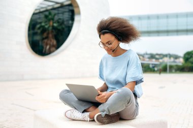 Focused african american curly haired young woman, with headset, consultant, mentor, in stylish formal clothes, sit with laptop outdoors while online work or study, looking for information in internet